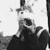 A person wears a whimsical animal hat and holds a Nikon camera up to their face. The scene is in black and white, with the background slightly blurred, creating an artistic and playful atmosphere. The person appears to be outdoors, possibly in a park or forested area.