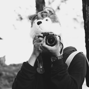 A person wears a whimsical animal hat and holds a Nikon camera up to their face. The scene is in black and white, with the background slightly blurred, creating an artistic and playful atmosphere. The person appears to be outdoors, possibly in a park or forested area.