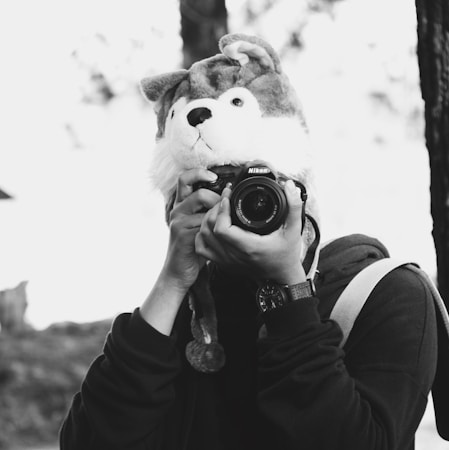 A person wears a whimsical animal hat and holds a Nikon camera up to their face. The scene is in black and white, with the background slightly blurred, creating an artistic and playful atmosphere. The person appears to be outdoors, possibly in a park or forested area.