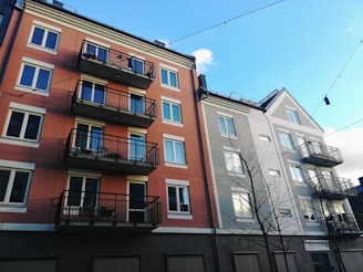 An urban street scene featuring two multi-story residential buildings with balconies. The building on the left has a warm orange facade, while the building on the right is painted in a light beige color with a peaked roof. There are several small balconies with black railings, and the sky above is clear with a few clouds.