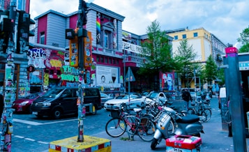 A vibrant urban scene featuring a street corner filled with buildings covered in colorful graffiti and street art. Numerous bicycles are parked alongside a row of cars. Trees line the streets, adding greenery amidst the concrete. People are seen casually walking and interacting, giving life to the busy street environment.