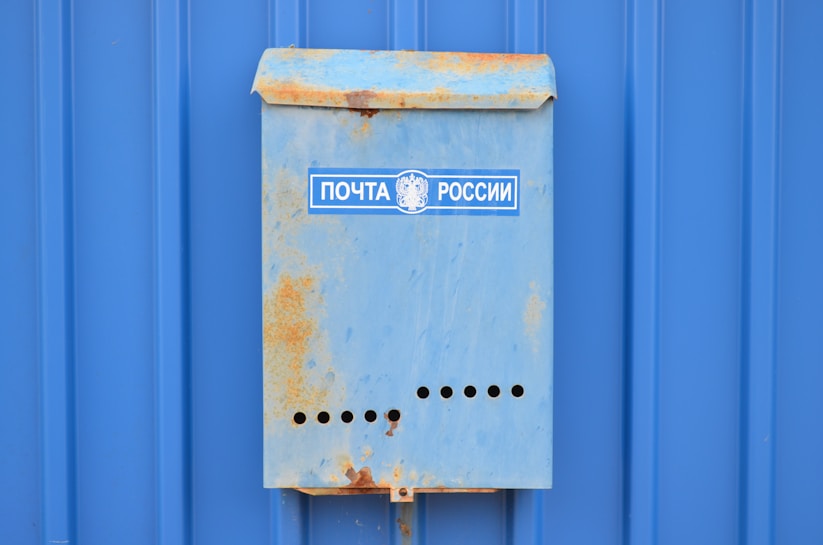 A weathered blue mailbox with rust spots is mounted on a blue corrugated metal wall. The mailbox has a label with Cyrillic text that suggests it is Russian.