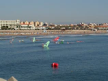 A beach with numerous people enjoying various water activities like windsurfing. Colorful sails are visible on the water as people engage in sports. The sandy beach is populated with sunbathers and umbrellas. In the background, there is a line of buildings with a clear blue sky above.