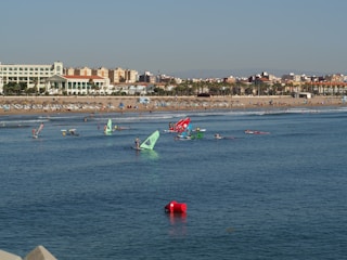 A beach with numerous people enjoying various water activities like windsurfing. Colorful sails are visible on the water as people engage in sports. The sandy beach is populated with sunbathers and umbrellas. In the background, there is a line of buildings with a clear blue sky above.