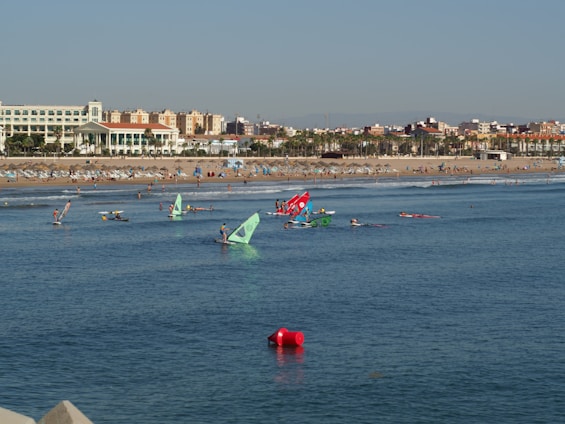 A beach with numerous people enjoying various water activities like windsurfing. Colorful sails are visible on the water as people engage in sports. The sandy beach is populated with sunbathers and umbrellas. In the background, there is a line of buildings with a clear blue sky above.
