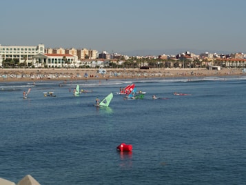 A beach with numerous people enjoying various water activities like windsurfing. Colorful sails are visible on the water as people engage in sports. The sandy beach is populated with sunbathers and umbrellas. In the background, there is a line of buildings with a clear blue sky above.