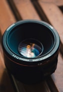 A camera lens resting on a wooden surface, featuring reflective glass showing intricate light patterns. The focus is on the text around the lens rim and the metallic details.
