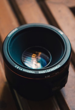 A camera lens resting on a wooden surface, featuring reflective glass showing intricate light patterns. The focus is on the text around the lens rim and the metallic details.