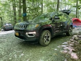A dark-colored SUV is parked in a forested campsite with other vehicles and camping equipment nearby. The ground is covered in gravel, and the area is surrounded by trees with green foliage. A red tent and a blue camping chair are visible in the background.
