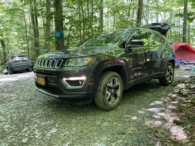 A dark-colored SUV is parked in a forested campsite with other vehicles and camping equipment nearby. The ground is covered in gravel, and the area is surrounded by trees with green foliage. A red tent and a blue camping chair are visible in the background.