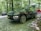A dark-colored SUV is parked in a forested campsite with other vehicles and camping equipment nearby. The ground is covered in gravel, and the area is surrounded by trees with green foliage. A red tent and a blue camping chair are visible in the background.