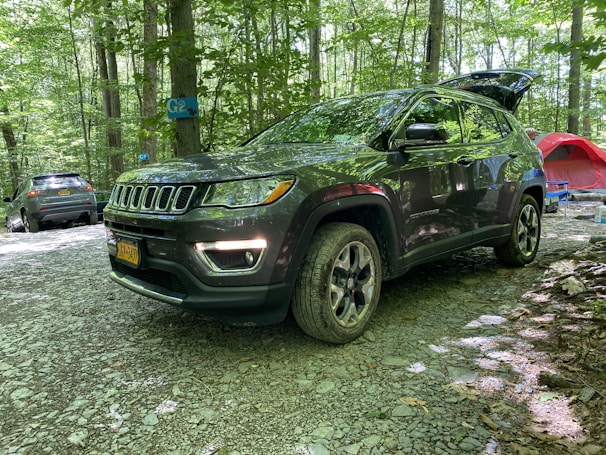 A dark-colored SUV is parked in a forested campsite with other vehicles and camping equipment nearby. The ground is covered in gravel, and the area is surrounded by trees with green foliage. A red tent and a blue camping chair are visible in the background.