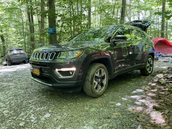 A dark-colored SUV is parked in a forested campsite with other vehicles and camping equipment nearby. The ground is covered in gravel, and the area is surrounded by trees with green foliage. A red tent and a blue camping chair are visible in the background.