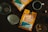 Two bright orange bags of Oak & Bond Coffee Co. are displayed on a wooden table. A cup of coffee, a metal coffee dripper, a paper filter, and a bottle with a cork are arranged around them, along with scattered coffee beans.