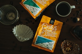 Two bright orange bags of Oak & Bond Coffee Co. are displayed on a wooden table. A cup of coffee, a metal coffee dripper, a paper filter, and a bottle with a cork are arranged around them, along with scattered coffee beans.