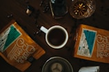 A wooden table with a cup of coffee at the center surrounded by two bags of coffee beans labeled Columbia and a glass with a dark liquid. Scattered coffee beans and a bottle stopper are spread around on the table.