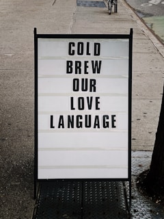 A sidewalk sign is displayed with a message written in bold black letters on a white background. The sign reads 'COLD BREW OUR LOVE LANGUAGE' and is set in an urban environment with a concrete sidewalk and a glimpse of a parked bicycle in the background.