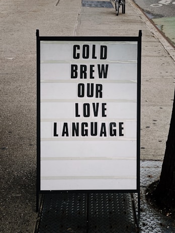 A sidewalk sign is displayed with a message written in bold black letters on a white background. The sign reads 'COLD BREW OUR LOVE LANGUAGE' and is set in an urban environment with a concrete sidewalk and a glimpse of a parked bicycle in the background.