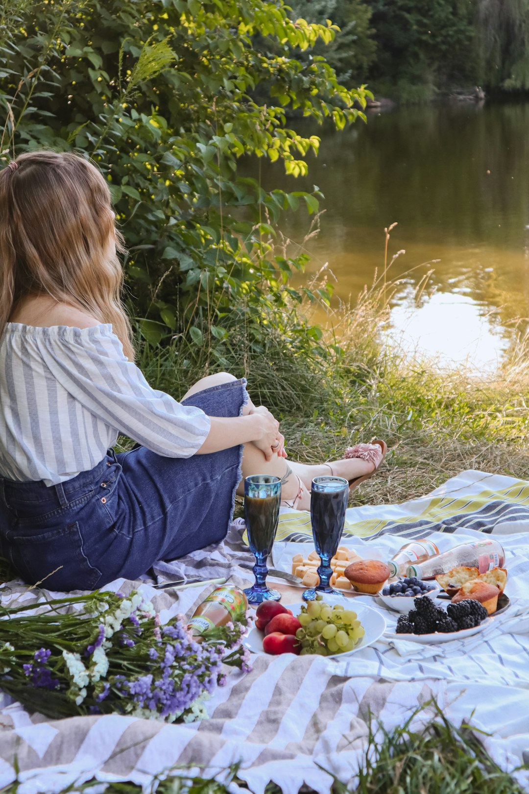 woman in white shirt and blue denim jeans sitting on green grass near lake during daytime