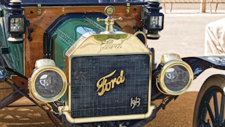Close-up view of the front of a vintage Ford car showcasing its brass radiator grille, prominent Ford logo, and rounded headlights. The car features polished metal and wooden elements, adding to its antique charm.