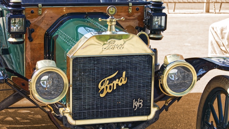 Close-up view of the front of a vintage Ford car showcasing its brass radiator grille, prominent Ford logo, and rounded headlights. The car features polished metal and wooden elements, adding to its antique charm.