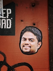 A sticker of a smiling man's face is placed on a textured, rusty orange surface. Above the sticker, part of a black and white printed text is visible, with the letters 'EP DAD'. The background has a rough, industrial look with visible bolt holes and faded black paint strokes across the bottom left corner.