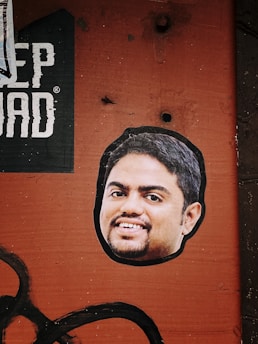 A sticker of a smiling man's face is placed on a textured, rusty orange surface. Above the sticker, part of a black and white printed text is visible, with the letters 'EP DAD'. The background has a rough, industrial look with visible bolt holes and faded black paint strokes across the bottom left corner.