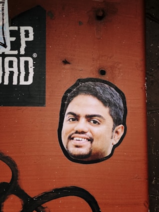 A sticker of a smiling man's face is placed on a textured, rusty orange surface. Above the sticker, part of a black and white printed text is visible, with the letters 'EP DAD'. The background has a rough, industrial look with visible bolt holes and faded black paint strokes across the bottom left corner.
