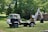 A vintage vehicle equipped with a backhoe attachment is parked on a grassy field. The truck has a weathered appearance with visible rust and wear, surrounded by lush green trees and foliage in the background. A small, older looking building is visible on the right.