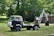 A vintage vehicle equipped with a backhoe attachment is parked on a grassy field. The truck has a weathered appearance with visible rust and wear, surrounded by lush green trees and foliage in the background. A small, older looking building is visible on the right.