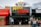 People are gathered outside of a movie theater called 'Movie Starr Cine-Plex'. The theater has a marquee with large yellow and red letters and is decorated with star-shaped lights. The surrounding area appears lively with a colorful red awning and other storefronts visible.
