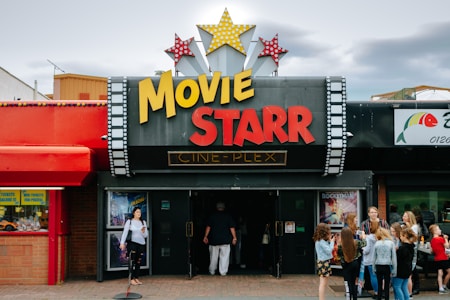 People are gathered outside of a movie theater called 'Movie Starr Cine-Plex'. The theater has a marquee with large yellow and red letters and is decorated with star-shaped lights. The surrounding area appears lively with a colorful red awning and other storefronts visible.