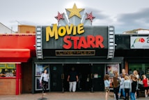 People are gathered outside of a movie theater called 'Movie Starr Cine-Plex'. The theater has a marquee with large yellow and red letters and is decorated with star-shaped lights. The surrounding area appears lively with a colorful red awning and other storefronts visible.