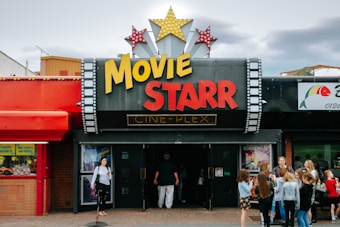 People are gathered outside of a movie theater called 'Movie Starr Cine-Plex'. The theater has a marquee with large yellow and red letters and is decorated with star-shaped lights. The surrounding area appears lively with a colorful red awning and other storefronts visible.