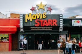 People are gathered outside of a movie theater called 'Movie Starr Cine-Plex'. The theater has a marquee with large yellow and red letters and is decorated with star-shaped lights. The surrounding area appears lively with a colorful red awning and other storefronts visible.