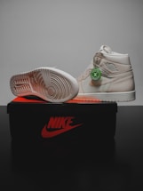 A pair of light beige sneakers are displayed on top of a black and orange Nike shoe box. One shoe is upright, showing the side and a green authenticity tag attached, while the other is positioned on its side to reveal the sole's tread pattern.