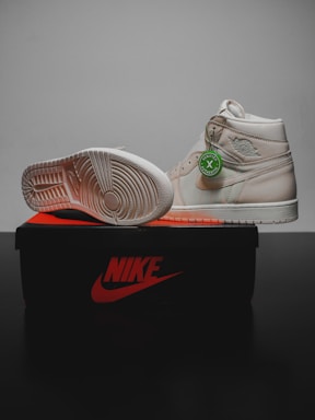 A pair of light beige sneakers are displayed on top of a black and orange Nike shoe box. One shoe is upright, showing the side and a green authenticity tag attached, while the other is positioned on its side to reveal the sole's tread pattern.