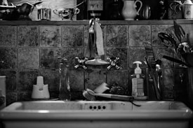 A monochrome kitchen scene features a rustic sink with well-used fixtures. A scrub brush, soap dispenser, and various kitchen utensils are visible around the sink area. Above the sink is a shelf holding various jars, a mortar and pestle, and a small pitcher, set against a backdrop of textured tiles that add an aged appearance.