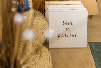 A collection of stacked wooden plaques featuring the phrase 'love is patient' written in elegant script. The setting includes large spools of twine and decorative sticks on a tiled floor, hinting at a rustic or craft-themed environment.