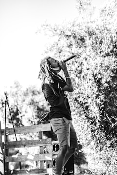 A person is performing outdoors, holding a microphone with their right hand. The scene captures the dynamic expression of their performance. They are wearing a t-shirt and shorts, with long hair styled in dreadlocks. The background features foliage and a wooden structure, emphasizing the outdoor setting.