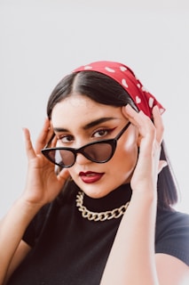 A person wears stylish black sunglasses and a red bandana with white polka dots. The makeup includes bold eyeliner and dark lipstick. The outfit features a black top and a chunky gold chain necklace. The individual holds the sunglasses with both hands, posing confidently.