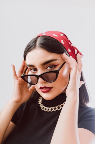 A person wears stylish black sunglasses and a red bandana with white polka dots. The makeup includes bold eyeliner and dark lipstick. The outfit features a black top and a chunky gold chain necklace. The individual holds the sunglasses with both hands, posing confidently.