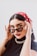 A person wears stylish black sunglasses and a red bandana with white polka dots. The makeup includes bold eyeliner and dark lipstick. The outfit features a black top and a chunky gold chain necklace. The individual holds the sunglasses with both hands, posing confidently.