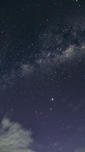 A star-filled night sky with a visible section of the Milky Way galaxy stretching diagonally across the image. There's a noticeable bright star centrally located on the lower half of the image, with some light clouds appearing in the bottom left corner.