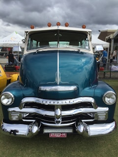 A classic blue Chevrolet truck is prominently displayed, featuring a shiny chrome grille and headlight design. The vehicle is parked on a grassy area, with a cloudy sky in the background and some event tents and people visible.
