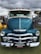 A classic blue Chevrolet truck is prominently displayed, featuring a shiny chrome grille and headlight design. The vehicle is parked on a grassy area, with a cloudy sky in the background and some event tents and people visible.