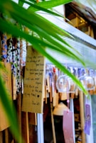 A collection of decorative wooden plaques and glass wind chimes hang together. One plaque bears a handwritten message with a heartfelt wish framed by natural green leaves providing a vibrant foreground. The scene exudes a crafted, artistic feel with various ribbons and strings adding to the visual interest.