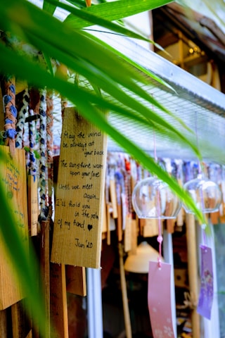 A collection of decorative wooden plaques and glass wind chimes hang together. One plaque bears a handwritten message with a heartfelt wish framed by natural green leaves providing a vibrant foreground. The scene exudes a crafted, artistic feel with various ribbons and strings adding to the visual interest.