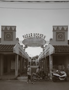 An entrance to a market labeled 'Pasar Modern' with architectural details typical of traditional buildings. Two decorative towers flank a central archway. A person is walking, and another is sitting on a motorbike next to a scooter. The scene is mostly empty with closed shops.