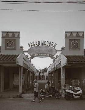 An entrance to a market labeled 'Pasar Modern' with architectural details typical of traditional buildings. Two decorative towers flank a central archway. A person is walking, and another is sitting on a motorbike next to a scooter. The scene is mostly empty with closed shops.